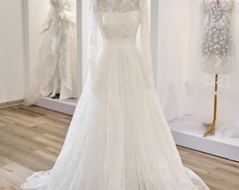 May include: A white wedding dress with long sleeves and a high neckline, displayed on a mannequin. The dress features intricate lace detailing and a flowing skirt. A gold crown sits atop the mannequin's head. Other white dresses are visible in the background.
