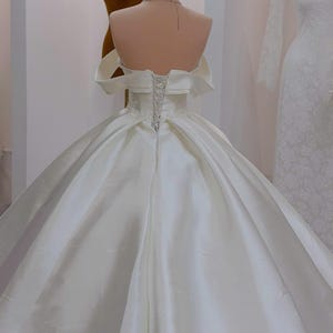 K&ouml;nnte beinhalten: Wei&szlig;es Brautkleid aus Satin mit schulterfreiem Ausschnitt und weitem Rock. Der R&uuml;cken hat einen Korsettverschluss. Eine goldene Krone sitzt auf einem Schaufensterpuppenkopf im Hintergrund, was auf Brautmode hindeutet.