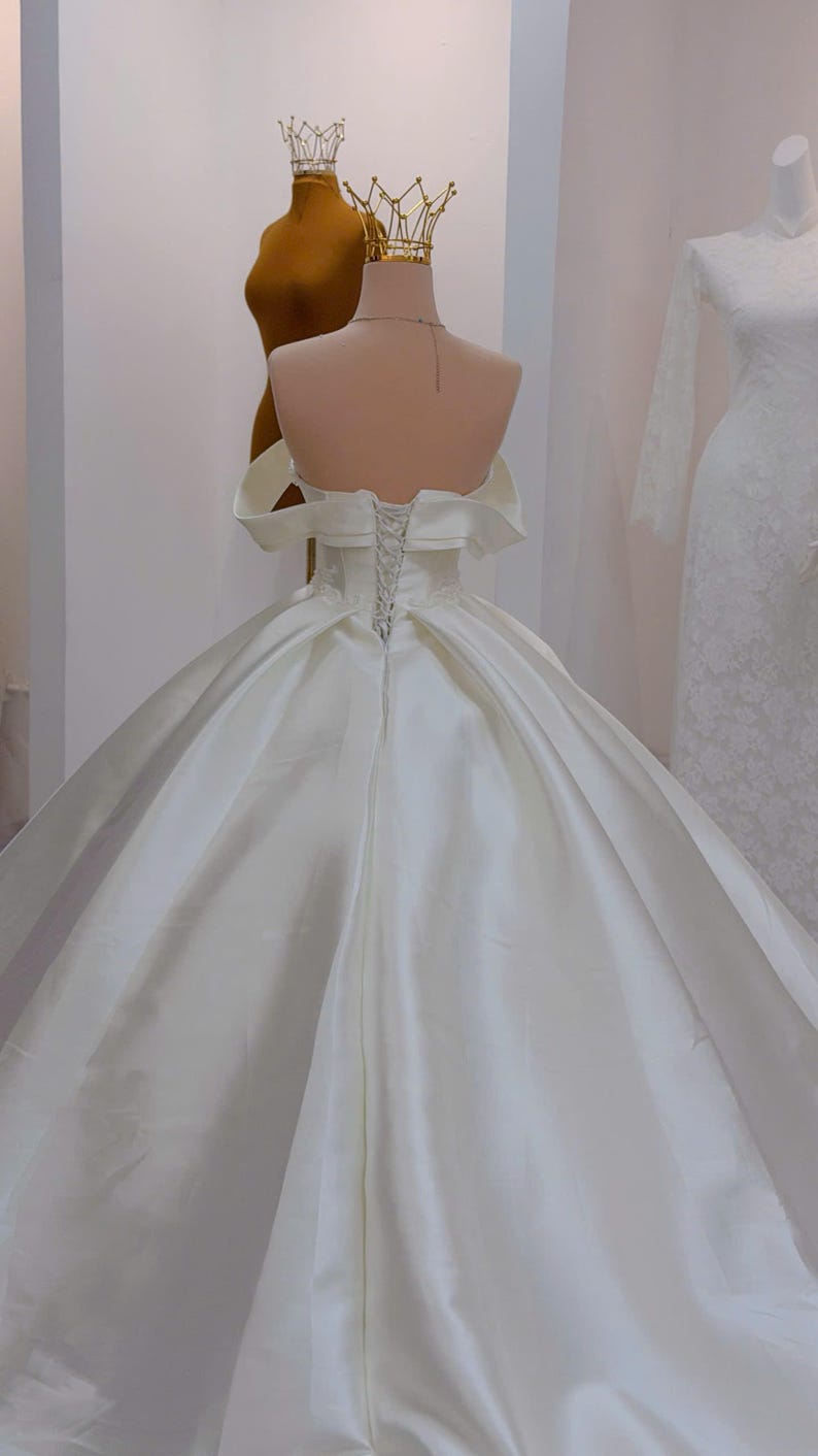 K&ouml;nnte beinhalten: Wei&szlig;es Brautkleid aus Satin mit schulterfreiem Ausschnitt und weitem Rock. Der R&uuml;cken hat einen Korsettverschluss. Eine goldene Krone sitzt auf einem Schaufensterpuppenkopf im Hintergrund, was auf Brautmode hindeutet.