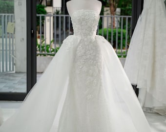 Puede incluir: Un vestido de novia blanco sin tirantes con bordado floral y cola desmontable se exhibe en un maniquí. Una corona dorada está colocada sobre la cabeza del maniquí. Otro vestido blanco cuelga en el fondo.
