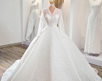 May include: A white lace wedding dress with long sleeves and a full skirt is displayed on a mannequin. The dress features a V-neckline and intricate floral lace detailing. A gold crown sits atop the mannequin's head, and other wedding dresses are visible in the background.