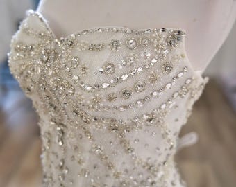 May include: A strapless white wedding dress bodice on a mannequin, heavily embellished with silver beads, sequins, and rhinestones. The intricate beadwork creates a sparkling, textured effect, ideal for bridal wear.