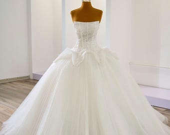 Puede incluir: Un vestido de novia blanco sin tirantes con una falda amplia y un corpiño con cuentas. El vestido se exhibe en un maniquí con una pequeña corona. El vestido presenta capas de tul y una silueta fluida.