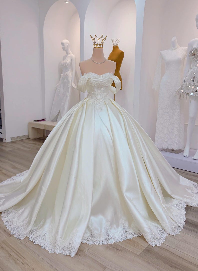 K&ouml;nnte beinhalten: Elfenbeinfarbenes Brautkleid aus Satin mit schulterfreiem Ausschnitt und weitem Rock. Das Kleid hat zarte Spitzendetails am Oberteil und am Saum. Eine goldene Krone sitzt auf dem Kopf der Schaufensterpuppe. Weitere wei&szlig;e Kleider sind im Hintergrund zu sehen.