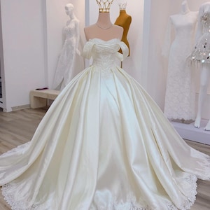 K&ouml;nnte beinhalten: Elfenbeinfarbenes Brautkleid aus Satin mit schulterfreiem Ausschnitt und weitem Rock. Das Oberteil hat zarte Spitzendetails, und der Rock geht in eine lange Schleppe mit Spitzenbesatz &uuml;ber. Eine goldene Krone sitzt auf dem Kopf der Schaufensterpuppe.