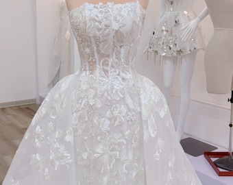 Puede incluir: Vestido de novia blanco sin tirantes con bordados florales y falda fluida. El vestido se exhibe en un maniquí, adornado con una corona dorada. Otros maniquíes con vestidos se ven en el fondo.