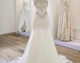 May include: A strapless white wedding gown with a mermaid silhouette. The bodice is embellished with sparkling crystals and beads. The dress is displayed on a mannequin, with a gold crown on its head. Other gowns are visible in the background.
