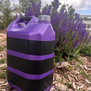 May include: A purple and black container with a handle and a grey cap. The container has three horizontal purple stripes on a black background. The container is on wood chips and grass, with purple flowers in the background.