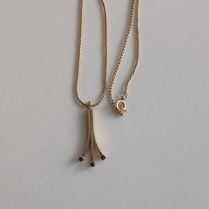 May include: A gold-colored necklace with a delicate chain and a pendant. The pendant features three downward-curving lines, each adorned with a small, square, red gemstone. The necklace is displayed on a white surface.