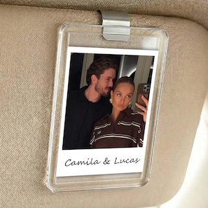 May include: A clear plastic car visor photo holder with a silver clip. The holder contains a photo of a couple, with the text "Camila & Lucas" printed at the bottom of the photo.