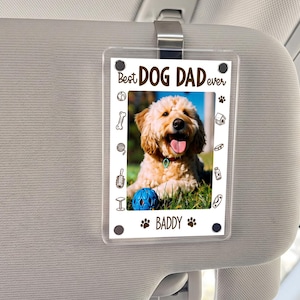 May include: A clear acrylic car visor accessory with the text "Best DOG DAD ever" and a photo of a dog. The photo is surrounded by a white border with dog-themed illustrations. The dog is light brown with a blue ball.