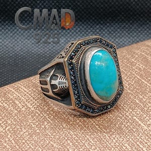 May include: A silver ring with a large, oval turquoise stone at the center, surrounded by small black stones. The ring has an ornate design with the text "CMAD 925" in the upper left corner.