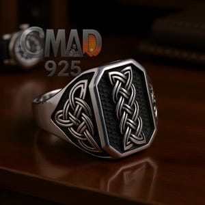 Puede incluir: Un anillo de plata con un diseño de nudo celta. El anillo tiene una cara rectangular con un fondo texturizado y un patrón de nudo detallado. El anillo está sobre una superficie de madera.