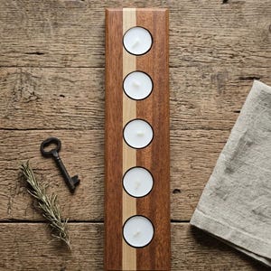 Puede incluir: Un portavelas de madera con cinco aberturas circulares para velas de té. El soporte es rectangular con madera marrón oscuro y una franja de madera clara en el centro. Una llave, una ramita de romero y una servilleta de lino están sobre una superficie de madera.