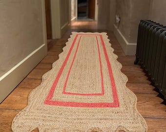 Puede incluir: Una alfombra rectangular de yute con borde festoneado y borde color coral. La alfombra está colocada sobre un suelo de madera en un pasillo. El color natural y el diseño de la alfombra añaden calidez y textura al espacio.