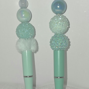 May include: Two decorative pens with mint green barrels and silver accents. The pens are topped with various decorative elements, including iridescent beads, glittery spheres, and a fluffy white accent. The pens are designed for writing.