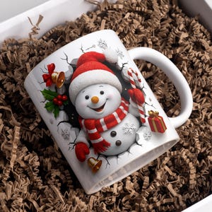 Puede incluir: Una taza de cerámica blanca con un diseño de muñeco de nieve en 3D. El muñeco de nieve lleva una bufanda, gorro y mitones rojos y blancos. La taza está decorada con copos de nieve, bastones de caramelo y una caja de regalo.