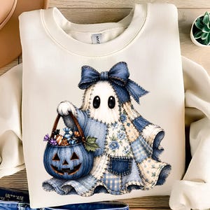 Puede incluir: Una sudadera de cuello redondo color crema con el gráfico de un fantasma que lleva una sábana estilo patchwork azul con un gran lazo a juego. El fantasma sostiene un cubo de calabaza azul lleno de caramelos envueltos de colores.
