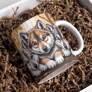 Może przedstawiać: Biały ceramiczny kubek z szczegółową ilustracją trzech szczeniąt wilka. Kubek ma biały uchwyt i jest umieszczony w pudełku z brązowym wypełniaczem papierowym. Wilki mają brązowe, szare i białe futro.