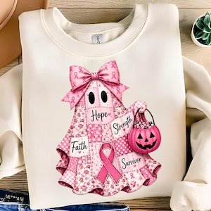 Pode incluir: Uma camisola de gola redonda cor de creme com um gráfico de um fantasma feito de tecido patchwork cor-de-rosa. O fantasma usa um grande laço cor-de-rosa, segura um balde de abóbora cor-de-rosa e exibe as palavras Faith, Hope, Strength e Survivor.