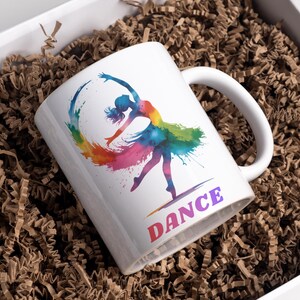 May include: White ceramic mug featuring a watercolor illustration of a ballerina in a rainbow-colored dress and a matching arc. The word "DANCE" is printed in colorful letters below the dancer. The mug is inside a white box filled with brown paper.