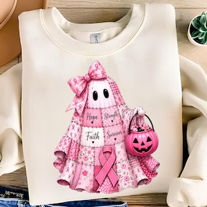 Op de afbeelding: Een crèmekleurig sweatshirt met ronde hals en een afbeelding van een geest in een roze patchwork-quiltpatroon. De geest draagt een grote roze strik, een lint voor borstkankerbewustzijn en houdt een roze pompoenemmer vast met de woorden Hope, Strength, Faith, Survivor.