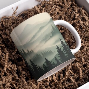 May include: White ceramic mug with a landscape design of a misty forest and mountains in green and grey shades. The mug is in a white box filled with brown crinkle paper.