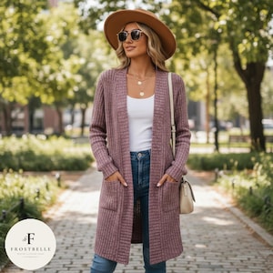 May include: A mauve-colored, long-sleeved cardigan with two front pockets. The open-front sweater is worn over a white top and blue jeans. The person is wearing a brown hat, sunglasses, and a cream-colored purse.