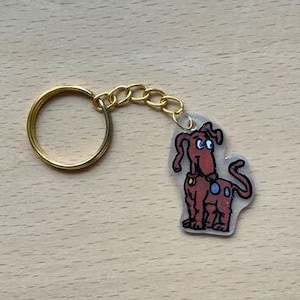Puede incluir: Un llavero novedoso con una ilustración de un perro de dibujos animados. El perro es marrón con manchas azules y un collar amarillo. El llavero tiene un anillo y una cadena dorados.