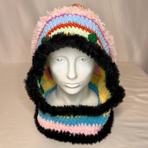 May include: A colorful, handmade crochet hood with a rainbow design. The hood features a pink, blue, and yellow color scheme with black faux fur trim around the face and neck. The hood is displayed on a white mannequin head.