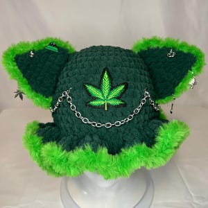 May include: Dark green crochet bucket hat with bright green faux fur trim. The hat features a green and black cannabis leaf patch and a silver chain. Small silver charms dangle from the ear flaps.