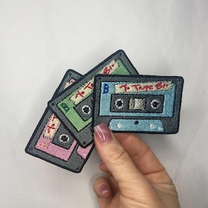 May include: Embroidered patches in the shape of retro cassette tapes. The patches are in shades of pink, green, and blue, with the text "Tape Bliss" visible on each. The patches have a gray border and are on a white background.