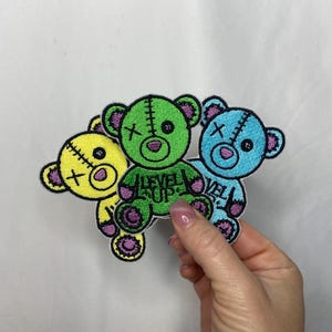 May include: Three embroidered teddy bear patches in yellow, green, and blue. Each bear has a stitched design, a cross on its face, and the text "LEVEL UP" on the green bear. The bears have pink accents and are held by a hand.