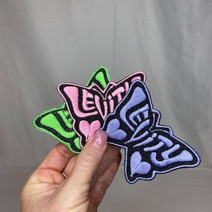 May include: Three embroidered butterfly patches in green, pink, and purple. Each butterfly has a black outline and the word "EVIL" in a stylized font. The patches are held in a hand against a white background.