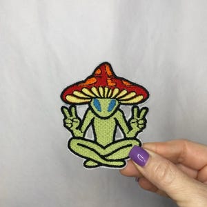 May include: Embroidered patch featuring a green alien with blue eyes, seated in a meditative pose, wearing a red and orange mushroom cap. The alien is making peace signs with both hands. The patch has a black border.