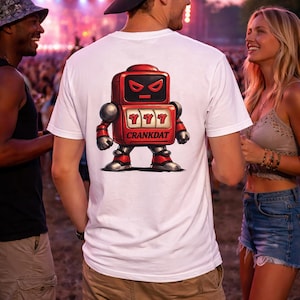 May include: White t-shirt featuring a red robot graphic with the text "CRANKDAT" and three sevens. The robot has a slot machine design. The image is taken at an outdoor event with people in the background.