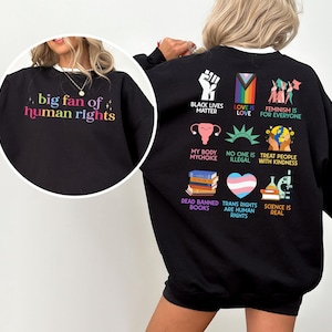 May include: Black sweatshirt with the text "big fan of human rights" in colorful letters on the front. The back of the sweatshirt features various graphics and phrases, including "Black Lives Matter" and "Love is Love."