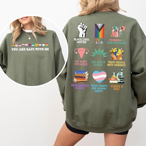 May include: An olive green sweatshirt with the text "YOU ARE SAFE WITH ME" and a rainbow of pride flags. The back features various graphics and phrases, including "BLACK LIVES MATTER," "LOVE IS LOVE," and "TRANS RIGHTS ARE HUMAN RIGHTS."