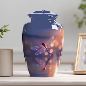 May include: A navy blue cremation urn with a dragonfly design. The urn has a glossy finish and a lid. The dragonfly is depicted with translucent wings and a purple body, set against a blurred sunset background.