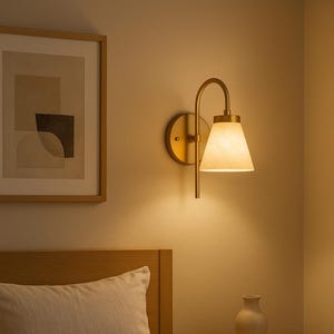 May include: A bedroom scene featuring a brass wall sconce with a conical shade, casting a warm glow. A framed abstract artwork hangs on the wall. The bed is made with neutral-toned linens, and a small vase sits on the nightstand.