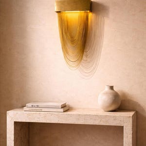 Könnte beinhalten: Moderne Wandleuchte mit rechteckiger Messingarmatur und goldfarbenem Kettenbehang. Die Leuchte ist an einer hellen Wand montiert und wirft einen Schatten. Eine kleine, cremefarbene Vase und zwei Bücher stehen auf einem Steintisch.