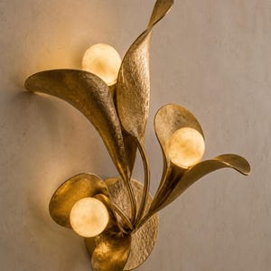 May include: A gold-toned wall sconce with a floral design. The sconce features three spherical, warm-toned lights nestled within stylized, textured metal leaves. The fixture is mounted on a neutral-colored wall.