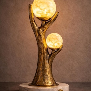 May include: A decorative table lamp with a bronze-coloured metal frame shaped like a tree. Two illuminated globe-shaped lights are held by the branches. The lamp sits on a round, marble-patterned base with a brass-coloured knob.