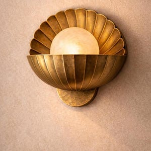 May include: A brass-toned wall sconce with a shell-like design. The fixture features a scalloped back and a bowl-shaped front, holding a frosted glass globe. The sconce is mounted on a circular base, creating a warm, inviting light.