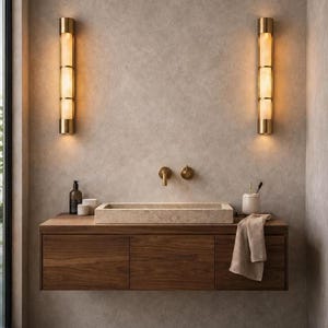May include: A modern bathroom vanity with a rectangular stone sink and a dark wood cabinet. Two vertical, gold-toned sconces flank the sink, providing warm lighting. A neutral-toned towel hangs on the counter, complementing the overall minimalist design.