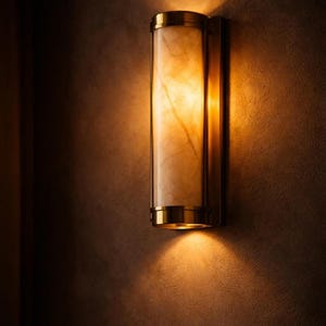 Puede incluir: Una lámpara de pared con una pantalla cilíndrica, similar al mármol, encerrada en un marco de latón. La luz emite un brillo cálido y acogedor, proyectando sombras en la pared texturizada. La lámpara está diseñada para uso en interiores.