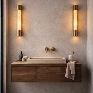 Peut inclure: Un meuble-lavabo suspendu en bois avec une vasque rectangulaire en pierre, un robinet mural en laiton et deux appliques murales cylindriques en laiton diffusant une lumière chaude. Une serviette beige est suspendue au meuble avec des accessoires.