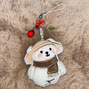 May include: A white plush bear charm wearing a brown scarf and hat. The bear has a black nose and eyes, with pink cheeks. The charm is attached to a silver clasp with a red bow, red bead, and green leaf accents.