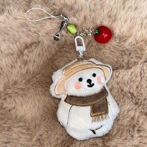 May include: A white plush bear charm with a tan hat and brown scarf, featuring pink cheeks and a black embroidered face. The charm includes a silver fork charm, green beads, and a red tomato charm. It is attached to a white string.