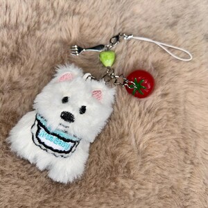 May include: A white plush dog charm with pink ears and a blue bib that says "KIDS STYLE". The charm has a silver fork, green apple, and red tomato charm attached. The charm is on a fluffy, tan surface.